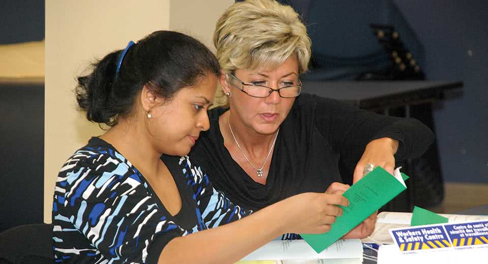 A woman with glasses teaches another woman from a WHSC workbook.